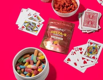 Delta-9 tablet pack with candy, playing cards, and snacks on a bright pink background