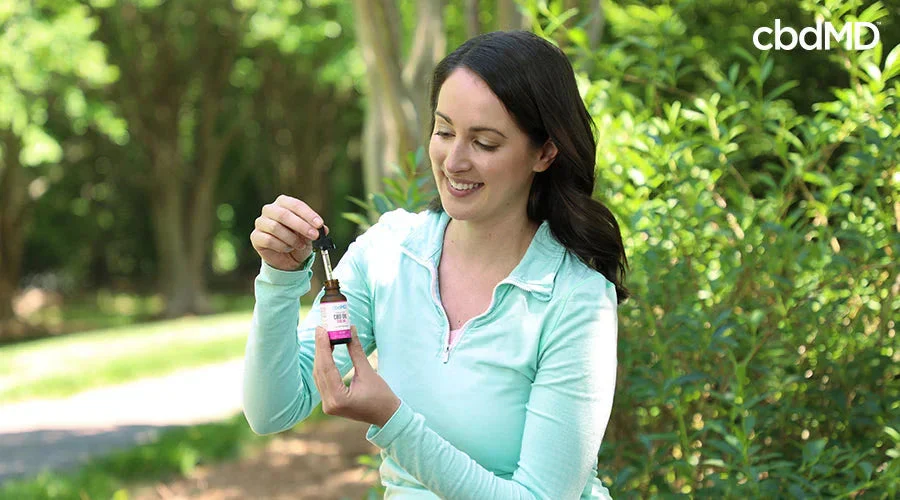 Woman outdoors holding CBD oil dropper bottle, smiling, with greenery in background