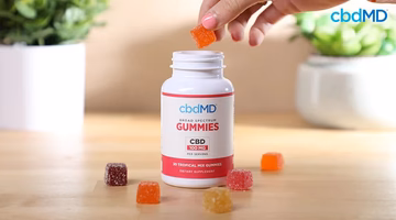 Hand taking a CBD gummy from an open bottle of assorted tropical CBD gummies on a table.