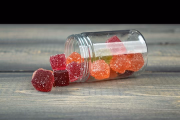 Assorted CBD gummies with sugar coating spilling from a clear jar onto a wooden surface