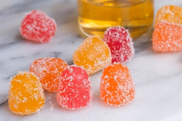 Assorted sugar-coated CBD gummies in bright colors on a marble surface with a glass jar.