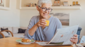 Smiling older woman with short gray hair reads newspaper and drinks orange juice at kitchen table