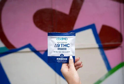 Hand holding hemp-derived Delta 9 THC gummies package, blue raspberry flavor, against colorful mural.