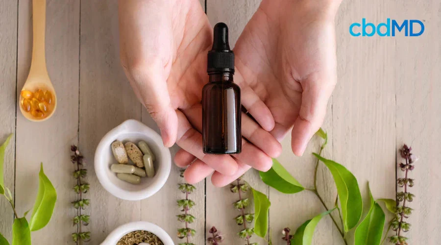 Hands holding a CBD oil dropper bottle with capsules, leaves, and herbs on a wooden surface