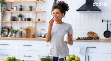 Woman in kitchen taking wellness supplement, holding bottle near fresh fruits and salad