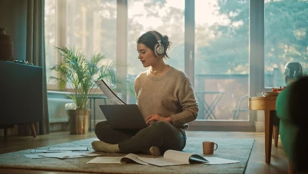 woman with headphones and a laptop in her lap sitting on the floor surrounded by papers