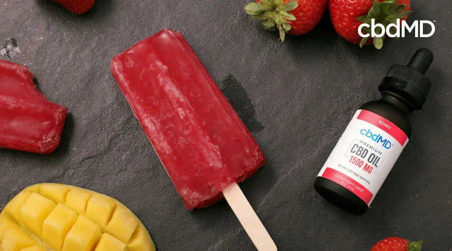 Berry CBD oil bottle with red popsicle, fresh strawberries, and sliced mango on slate surface