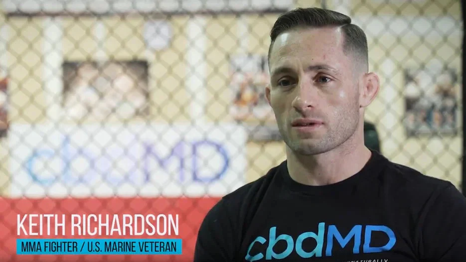 MMA fighter in black cbdMD shirt sitting in gym with chain-link fence background