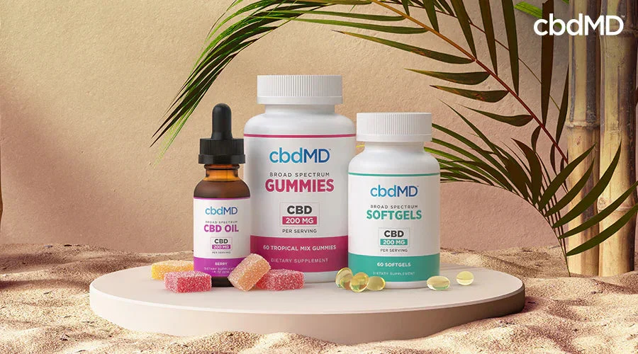 CBD oil dropper bottle, gummies, and softgels on a display with palm leaves and sand.