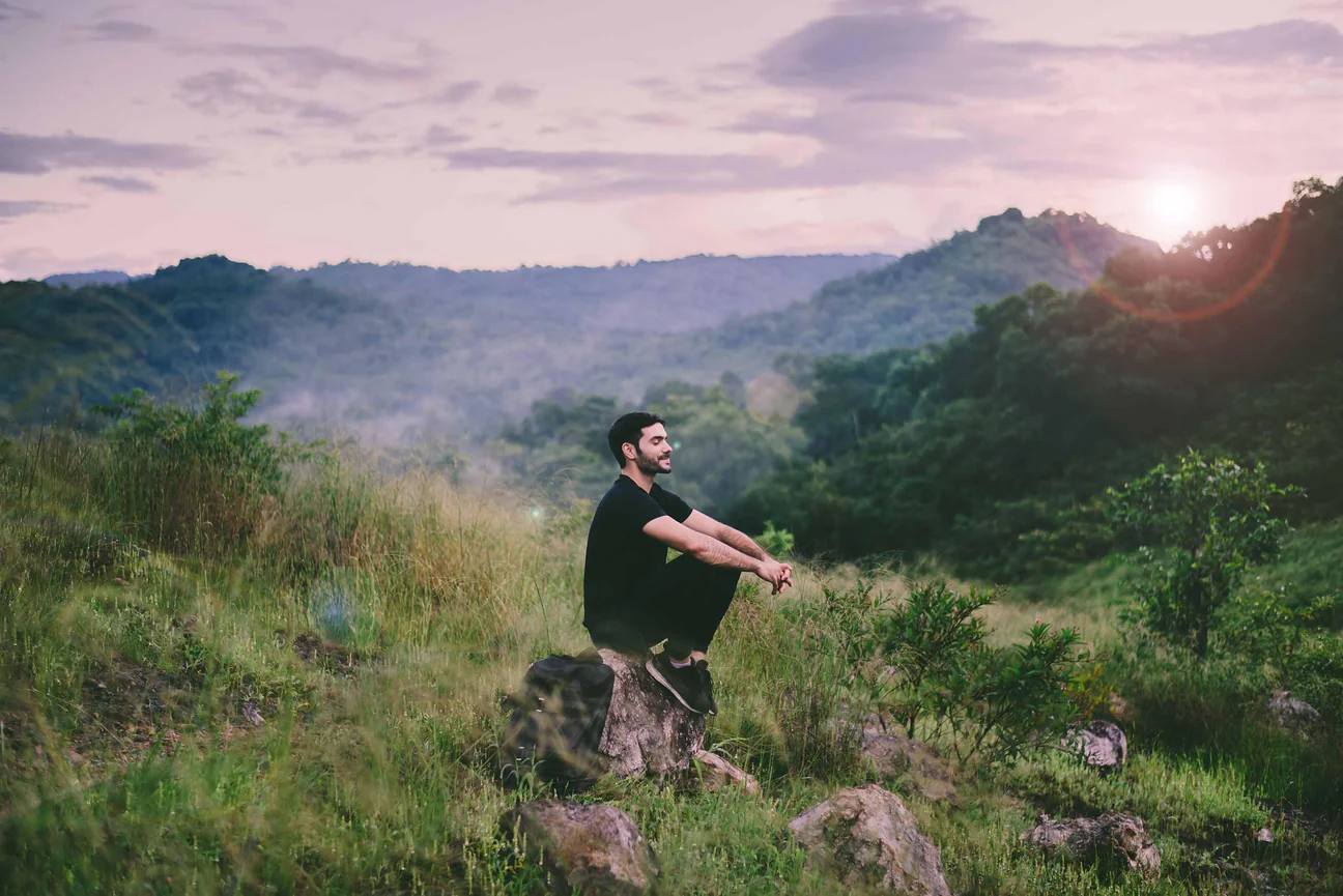 Man meditating on a grassy hillside at sunrise with peaceful mountain scenery