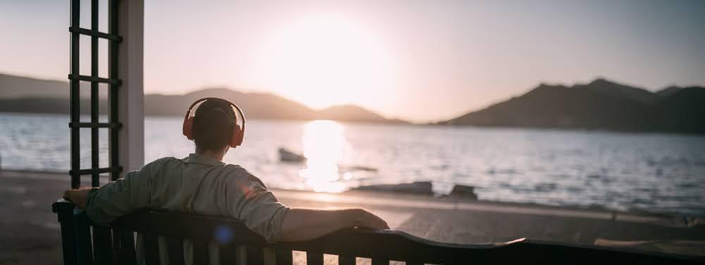Man with headphones relaxing on a bench by the water at sunrise, mountains in background