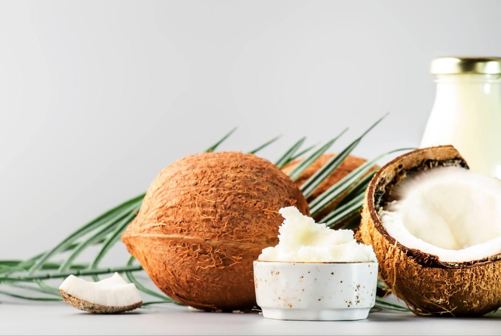 Whole coconuts, coconut oil in a jar, and coconut meat on a white table with palm leaves