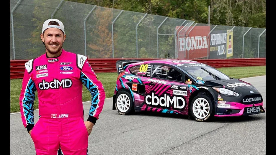 Race car driver in pink cbdMD suit standing beside branded rally car on racetrack