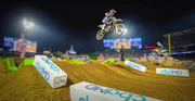 Motocross rider jumping dirt ramp at night supercross event with sponsor signs on track