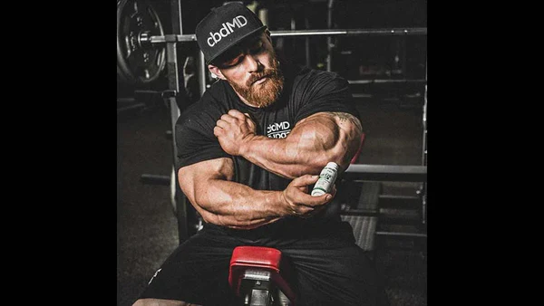 Muscular man in gym applying topical cream to arm, wearing cbdMD shirt and hat