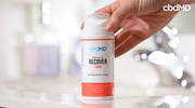 Hand holding a bottle of premium recover pain relieving cream in a bathroom setting.