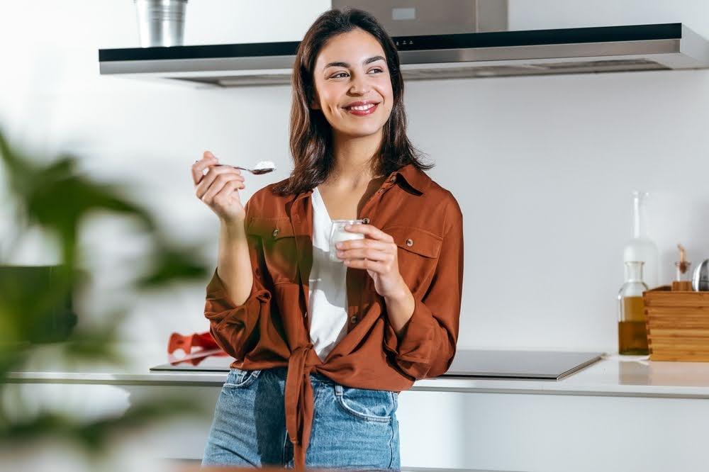 Smiling woman in casual clothes eating yogurt in a modern kitchen, healthy lifestyle concept.