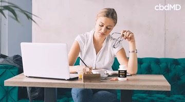Woman working on laptop at table with CBD gummies bottle, glasses, and notebook