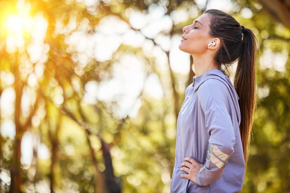 Woman runner in park enjoying sunlight, wearing athletic jacket and earbuds