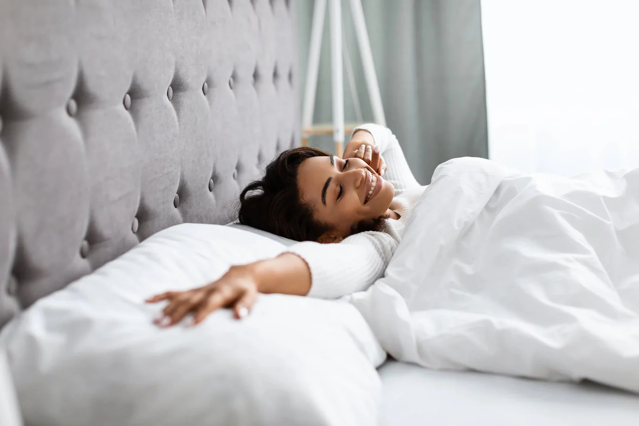 Smiling woman waking up and stretching in bed with white bedding and soft headboard