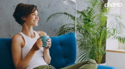 Smiling woman relaxing on blue sofa with cup of calming CBD tea and lush green plant nearby